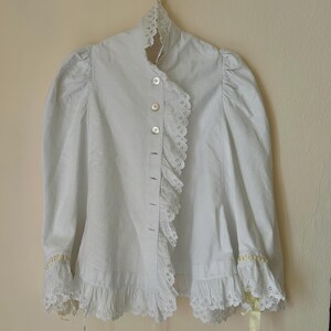 May include: A white blouse with a high collar and ruffled trim. The blouse features long, puffy sleeves and is buttoned down the front. Delicate lace and yellow ribbon accents adorn the cuffs. The garment hangs on a gold hanger.