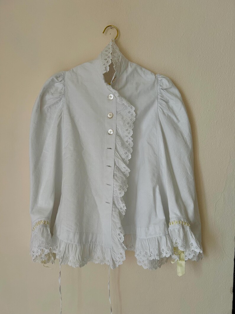 May include: A white blouse with a high collar and ruffled trim. The blouse features long, puffy sleeves and is buttoned down the front. Delicate lace and yellow ribbon accents adorn the cuffs. The garment hangs on a gold hanger.