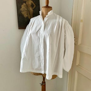 May include: A white long-sleeved blouse featuring a high, ruffled collar and a button-down front. The garment has voluminous sleeves with gathered shoulders and a straight hemline, displayed on a wooden dress form mannequin.