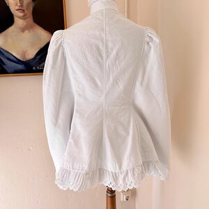 May include: A white, vintage-style jacket with a high collar and long sleeves. The jacket features a ruffled hem and delicate lace trim. The garment is displayed on a wooden mannequin, showcasing its tailored silhouette and intricate details.
