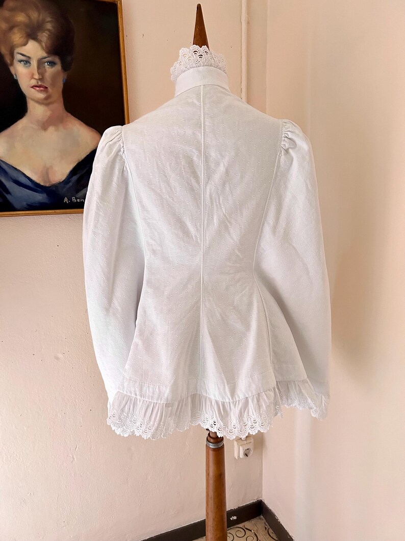 May include: A white, vintage-style jacket with a high collar and long sleeves. The jacket features a ruffled hem and delicate lace trim. The garment is displayed on a wooden mannequin, showcasing its tailored silhouette and intricate details.
