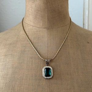 May include: A gold-toned necklace with a pendant featuring a large, emerald-green rectangular stone surrounded by small, clear crystals. The necklace is displayed on a tan mannequin.