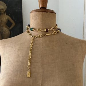 May include: A gold chain necklace with a long chain and a small tag that says "C.F." The necklace has a double strand of gold chain with green, blue, and red beads.