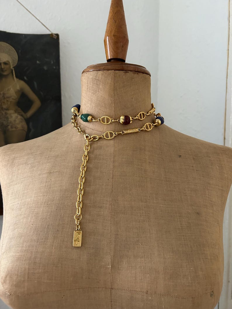May include: A gold chain necklace with a long chain and a small tag that says "C.F." The necklace has a double strand of gold chain with green, blue, and red beads.