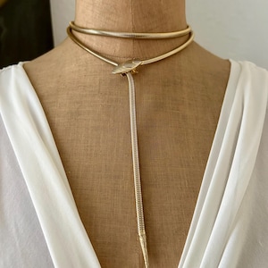 Op de afbeelding: Een goudkleurige slangenketting met een flexibel, buisvormig ontwerp dat om de nek wikkelt. De ketting bevat een centrale, ovaalvormige metalen sluiting die de ketting vastzet, waardoor een enkele streng verticaal naar beneden hangt.