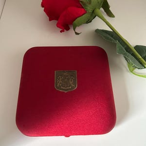 Op de afbeelding: Een rode fluwelen sieradendoos met een gouden embleem, naast een rode roos met groene bladeren. De doos is vierkant met afgeronde hoeken. Het embleem heeft de vorm van een schild met een gedetailleerd ontwerp. De roos staat in volle bloei.
