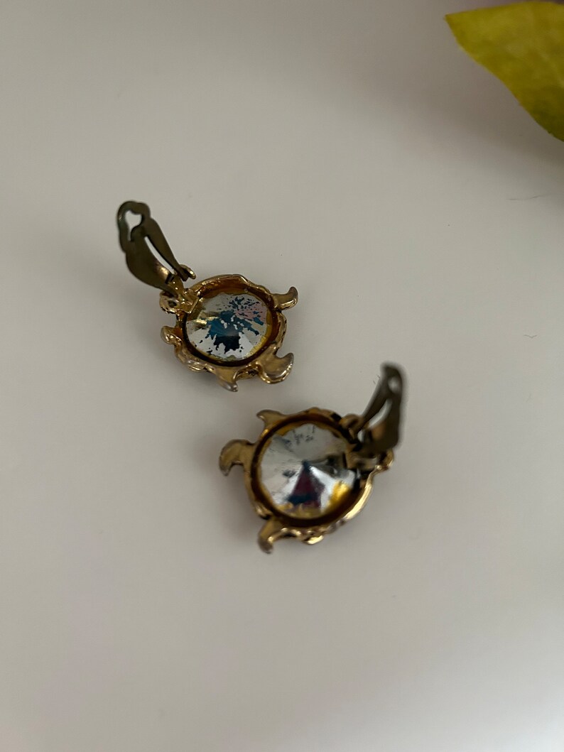 K&ouml;nnte beinhalten: Ein Paar Vintage-Ohrclips. Jeder Ohrring hat einen gro&szlig;en, klaren, facettierten Kristallstein, der in einen goldfarbenen Metallrahmen gefasst ist. Der Rahmen hat einen dekorativen, gewellten Rand und einen Clip-Mechanismus.