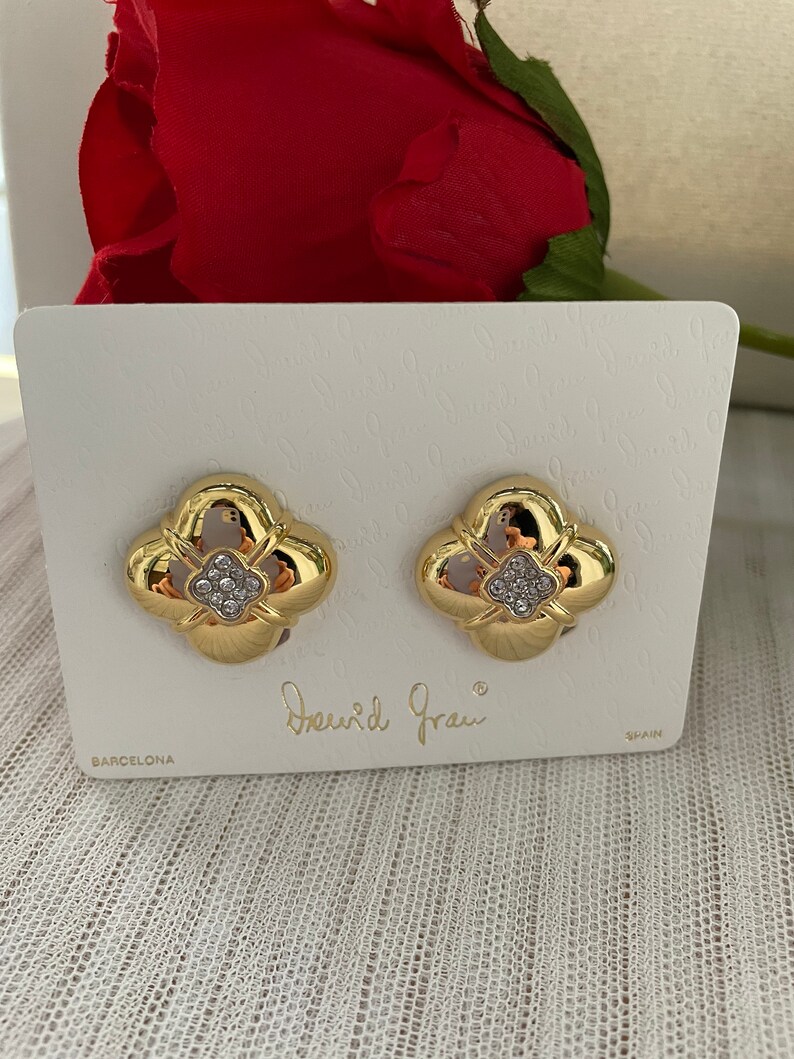 May include: Gold tone earrings with a four-leaf clover design and clear rhinestones. The earrings are on a white card with the text "David Grau" and "Barcelona" printed on it.