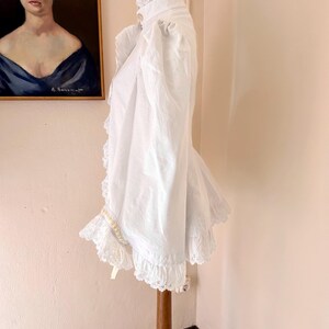 May include: A white vintage blouse with a high collar and ruffled trim. The blouse features lace detailing on the collar, cuffs, and hem. The garment is displayed on a wooden stand, showcasing its design and texture.