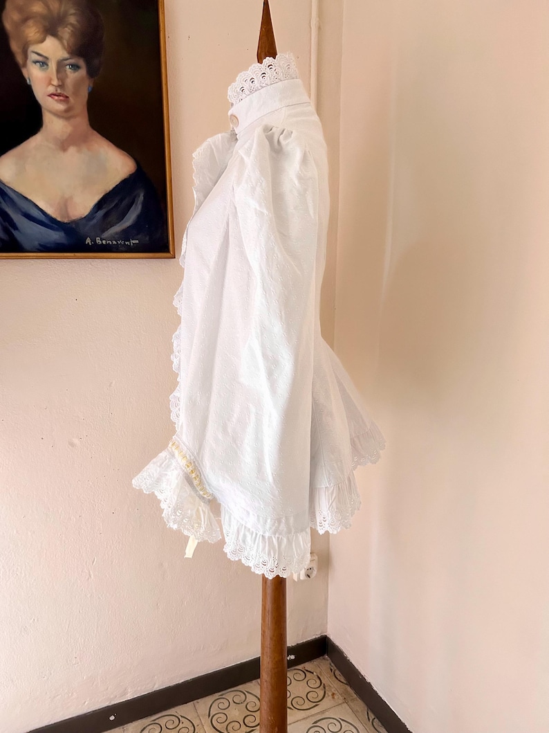 May include: A white vintage blouse with a high collar and ruffled trim. The blouse features lace detailing on the collar, cuffs, and hem. The garment is displayed on a wooden stand, showcasing its design and texture.