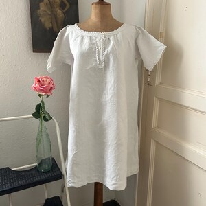 May include: A white linen nightgown with short sleeves and a round neckline, displayed on a wooden mannequin. The nightgown features delicate lace trim around the neckline and sleeves, with a small buttoned placket. A pink rose in a glass bottle is visible in the background.