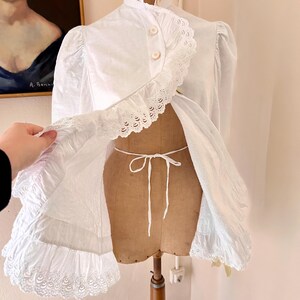 May include: White vintage jacket with a high collar, button closure, and ruffled lace trim. The jacket is displayed on a vintage dress form. The jacket has long sleeves and a tie closure at the waist. The jacket is made of a lightweight fabric.