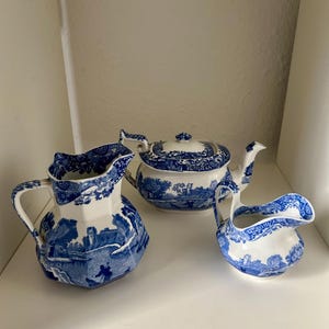 Puede incluir: Un juego de té de cerámica que consta de una jarra grande, una tetera con tapa y una lechera más pequeña. Cada pieza presenta una base blanca con un patrón azul detallado que representa un paisaje con edificios, árboles y figuras.
