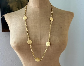 May include: A long gold-tone chain necklace featuring evenly spaced circular medallions. Each medallion displays a raised sun-like face design with a textured border. The necklace is displayed on a beige fabric-covered mannequin torso.