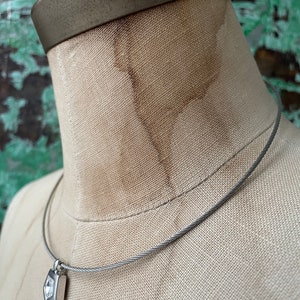 May include: A silver necklace with a small, square pendant featuring a clear, faceted gemstone. The necklace is made of a braided cable chain.