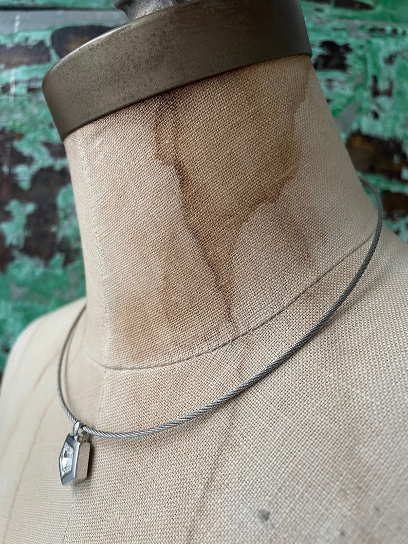 May include: A silver necklace with a small, square pendant featuring a clear, faceted gemstone. The necklace is made of a braided cable chain.