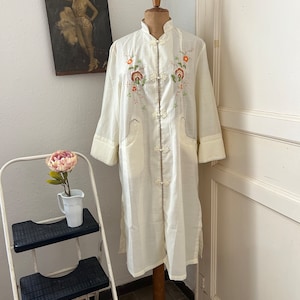 Puede incluir: Una bata de seda blanca con bordados florales y cuello mandarín. La bata tiene una cremallera y dos bolsillos.