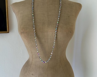 May include: A long, clear beaded necklace with iridescent accents hangs on a vintage dress form. The beads are small and round, reflecting a rainbow of colors. The necklace is a delicate piece of jewelry.