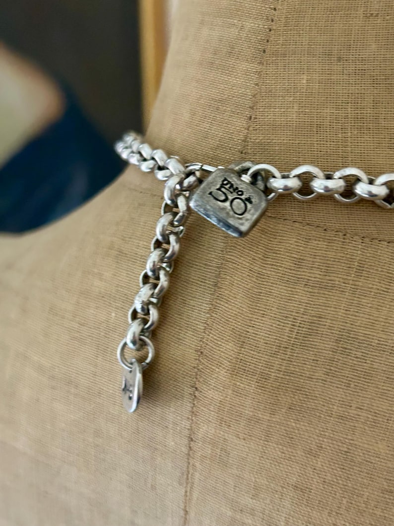 May include: Silver-toned necklace with a chunky chain design. The necklace features a square pendant with the number "50" engraved on it. The necklace is displayed on a mannequin.
