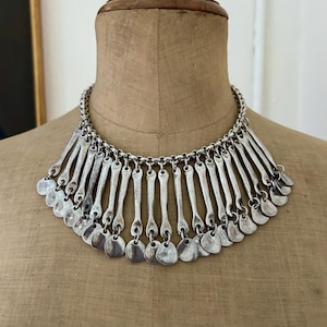 May include: Silver-toned statement necklace with a chain and pendant design. The necklace features a series of vertical bars and small, round pendants. The necklace is displayed on a mannequin.