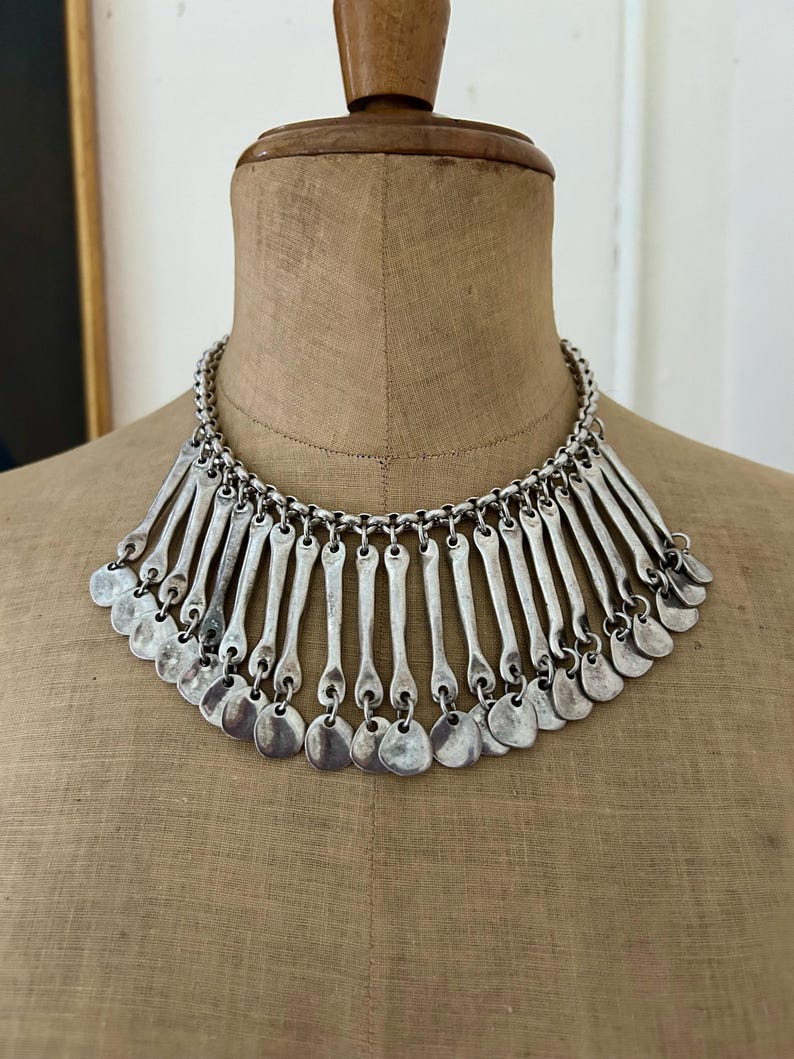 May include: Silver-toned statement necklace with a chain and pendant design. The necklace features a series of vertical bars and small, round pendants. The necklace is displayed on a mannequin.