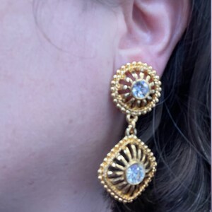May include: Gold tone dangle earrings with clear rhinestone accents. The earrings feature a round top with a smaller square dangle.