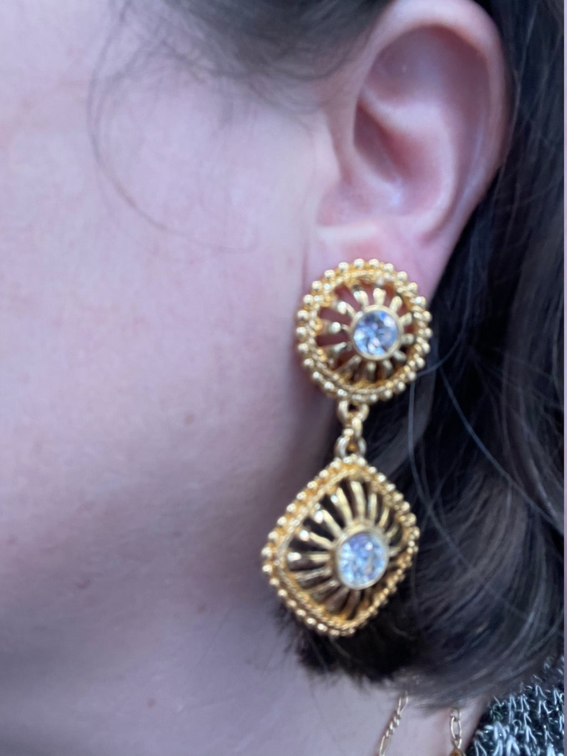 May include: Gold tone dangle earrings with clear rhinestone accents. The earrings feature a round top with a smaller square dangle.