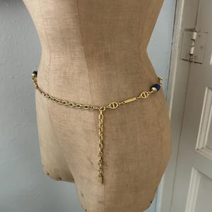 May include: A gold chain belt with blue oval-shaped beads. The chain has a small gold tag hanging from it.