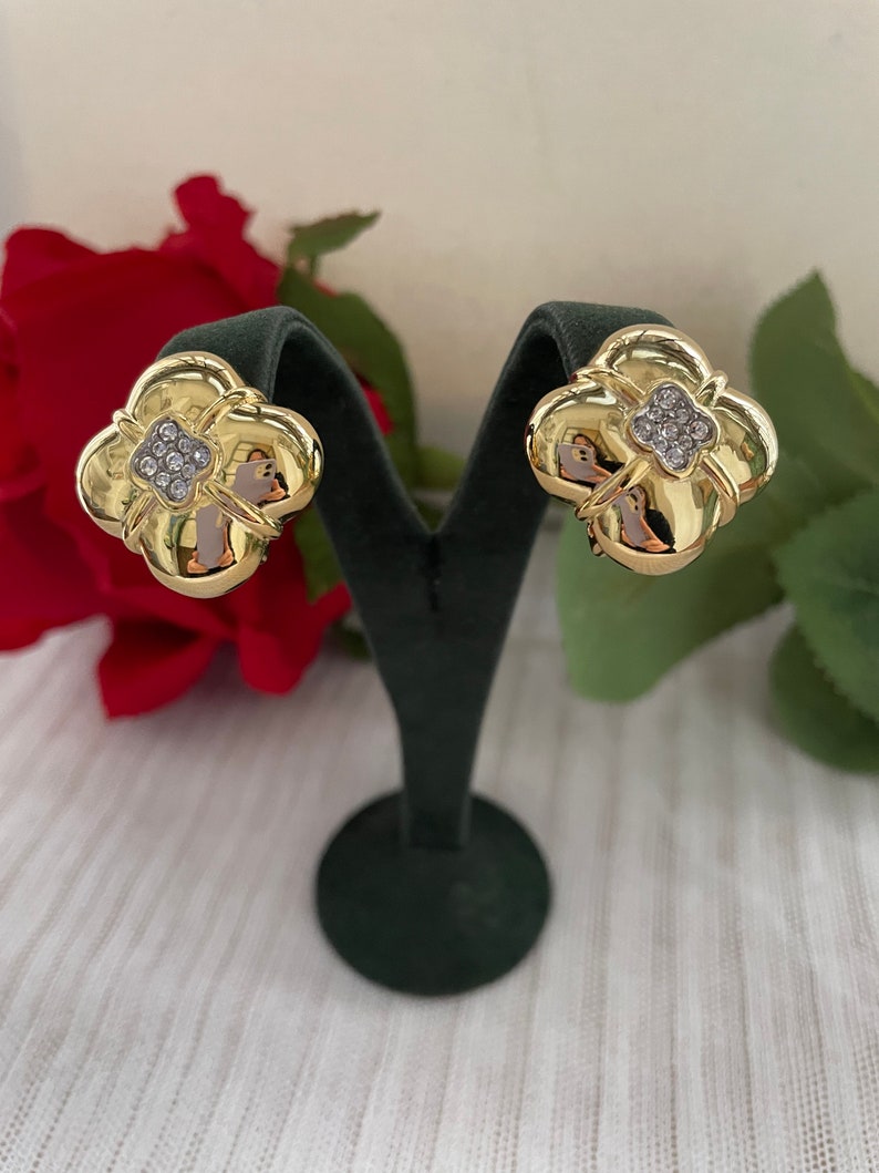 May include: A pair of gold-tone earrings with a four-leaf clover design. Each earring features a cluster of clear rhinestones in the center.