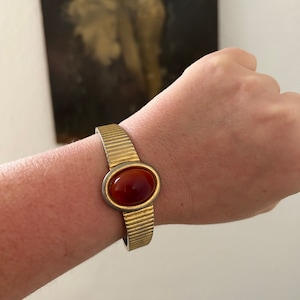 May include: A gold-toned bangle bracelet with a ribbed design and a large, oval-shaped, red-brown gemstone in the centre.