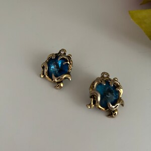 K&ouml;nnte beinhalten: Zwei verzierte Ohrringe mit goldfarbenem Filigranmuster. Jeder Ohrring hat einen runden, tiefblauen Stein und einen funkelnden Akzent. Sie liegen auf einem wei&szlig;en Hintergrund.