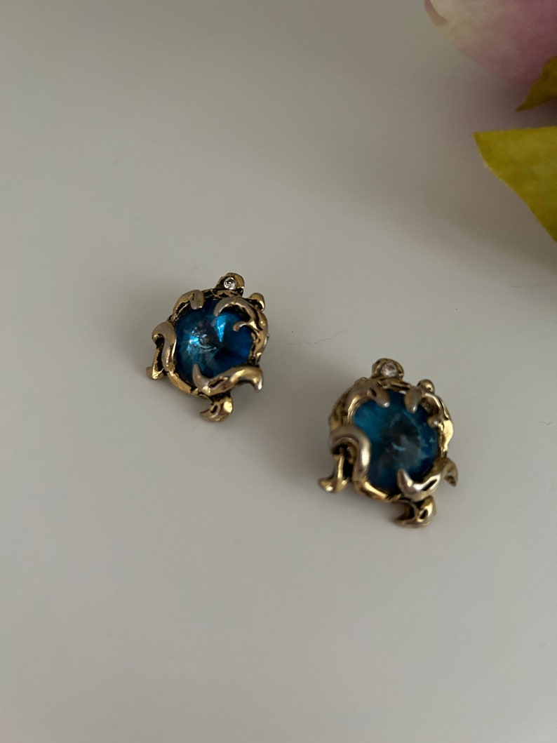 K&ouml;nnte beinhalten: Zwei verzierte Ohrringe mit goldfarbenem Filigranmuster. Jeder Ohrring hat einen runden, tiefblauen Stein und einen funkelnden Akzent. Sie liegen auf einem wei&szlig;en Hintergrund.