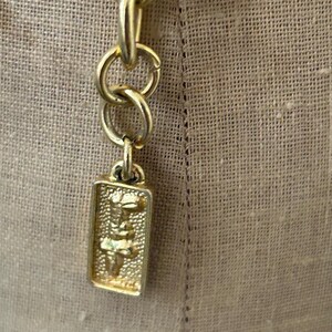 May include: A gold chain with a rectangular tag attached. The tag has a detailed design of a stylized figure.