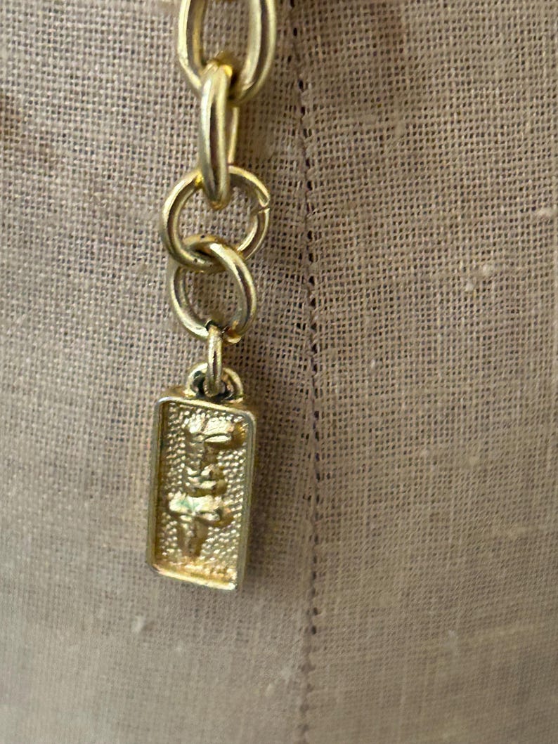 May include: A gold chain with a rectangular tag attached. The tag has a detailed design of a stylized figure.