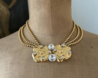 May include: A multi-strand gold-tone beaded necklace featuring a central decorative element composed of two polished gold-tone flower motifs accented with clear, round faceted crystals set between the floral shapes and at the center.
