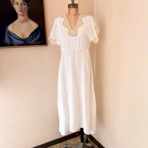 Puede incluir: Un vestido blanco vintage con mangas cortas y cuello en V, exhibido en un maniquí. El escote y los bordes de las mangas presentan un delicado encaje beige. El vestido es largo hasta el suelo y tiene un diseño simple y elegante. Una pintura cuelga en el fondo.
