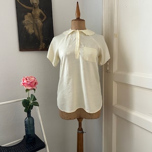 May include: A white cotton blouse with a Peter Pan collar and short sleeves. The blouse has a small pocket on the left side and is decorated with embroidered flowers.