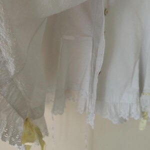 May include: White cotton garment with lace trim and yellow accents. The garment features a ruffled hem and delicate lace detailing. Buttons are visible on the front. The overall style is vintage and feminine.