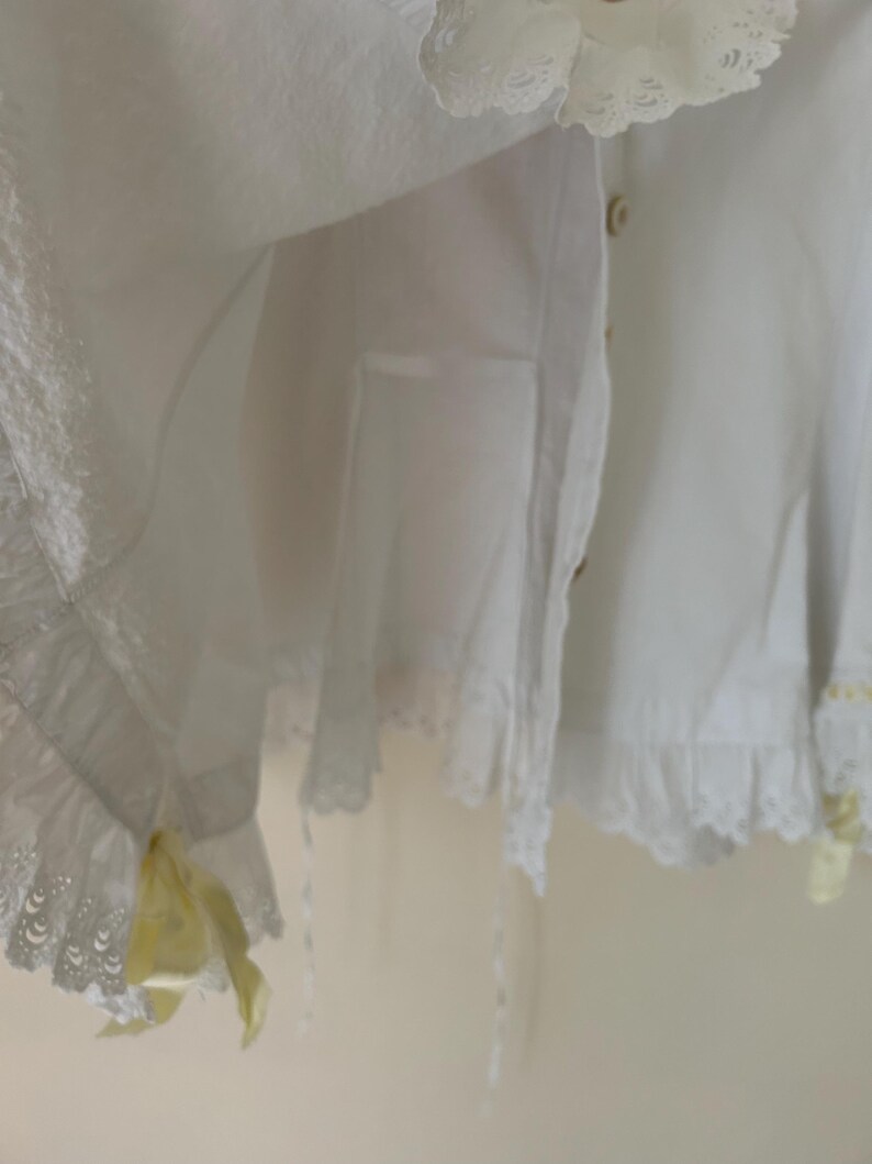 May include: White cotton garment with lace trim and yellow accents. The garment features a ruffled hem and delicate lace detailing. Buttons are visible on the front. The overall style is vintage and feminine.