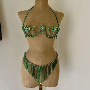 May include: A green rhinestone bra and matching bottom set displayed on a mannequin. The bra features floral accents and a halter neck. The bottom piece has a fringe design. The set is made of green rhinestones.