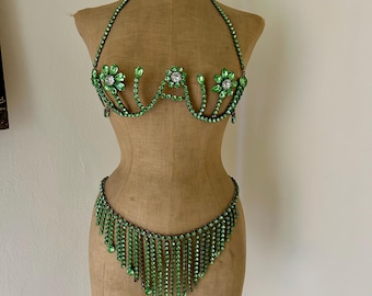 Könnte beinhalten: Ein grünes Strass-BH und passendes Unterteil-Set, das auf einer Schaufensterpuppe ausgestellt ist. Der BH hat florale Akzente und einen Neckholder. Das Unterteil hat ein Fransen-Design. Das Set besteht aus grünen Strasssteinen.