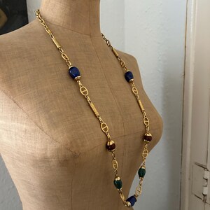 May include: A gold chain necklace with blue, green, and brown oval-shaped beads. The chain features a repeating interlocking 'D' design.