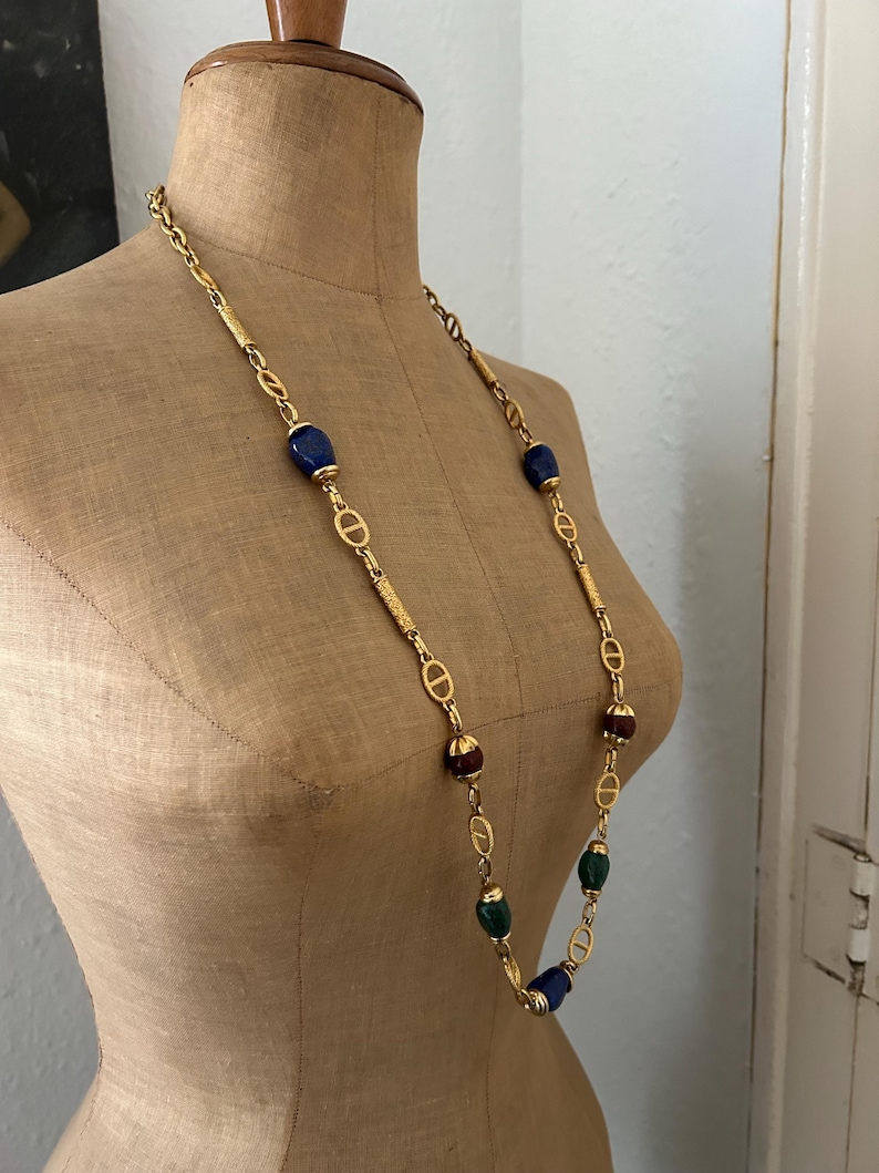 May include: A gold chain necklace with blue, green, and brown oval-shaped beads. The chain features a repeating interlocking 'D' design.