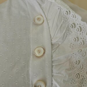 May include: Close-up of a white garment with three large, round buttons down the front. The fabric has a subtle diamond pattern on one side and a ruffled, embroidered edge on the other. The buttons are a creamy white color.