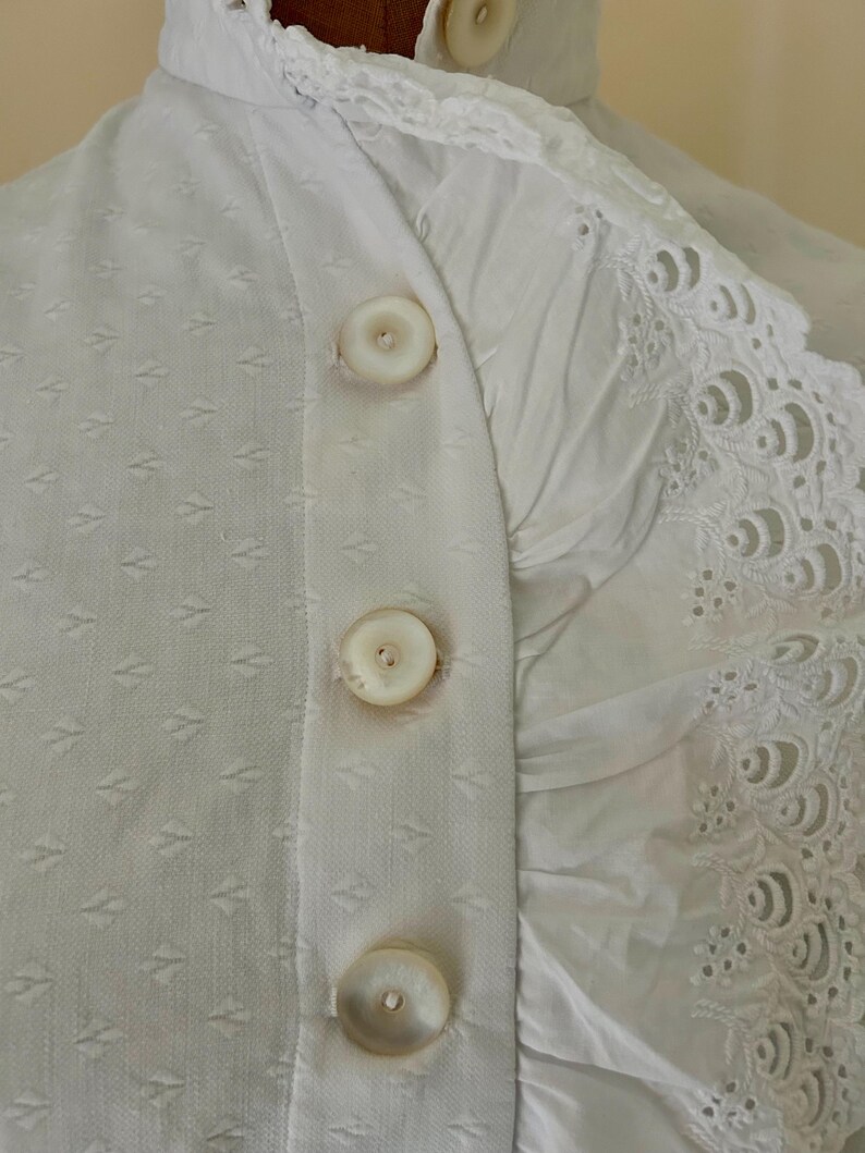May include: Close-up of a white garment with three large, round buttons down the front. The fabric has a subtle diamond pattern on one side and a ruffled, embroidered edge on the other. The buttons are a creamy white color.