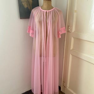 May include: A floor-length, sheer light pink robe featuring short, loose sleeves with matching pink lace trim at the cuffs. The garment has a round neckline with a single small button closure at the top and an open front design made of lightweight fabric.