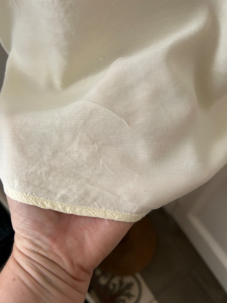 May include: Close-up of a cream-coloured fabric with a subtle texture. The fabric has a yellowed edge with a stitched seam.