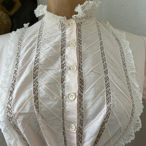 K&ouml;nnte beinhalten: Eine rosa Baumwollbluse mit wei&szlig;en Spitzenbesatz und Knopfleiste. Der Spitzenbesatz ist in einem Chevron-Muster.