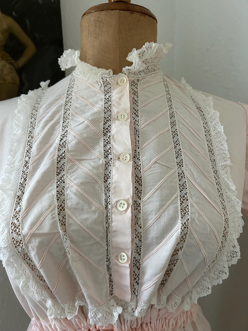 K&ouml;nnte beinhalten: Eine rosa Baumwollbluse mit wei&szlig;en Spitzenbesatz und Knopfleiste. Der Spitzenbesatz ist in einem Chevron-Muster.