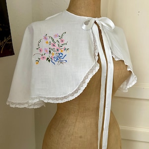 Könnte beinhalten: Ein weißes Capelet mit einem zarten Blumenstickmuster in Rosa, Gelb und Grün sowie einem blauen Schleifenakzent. Das Kleidungsstück ist am geschwungenen Saum mit weißer Spitze verziert und verfügt über ein weißes Satinband als Verschluss.